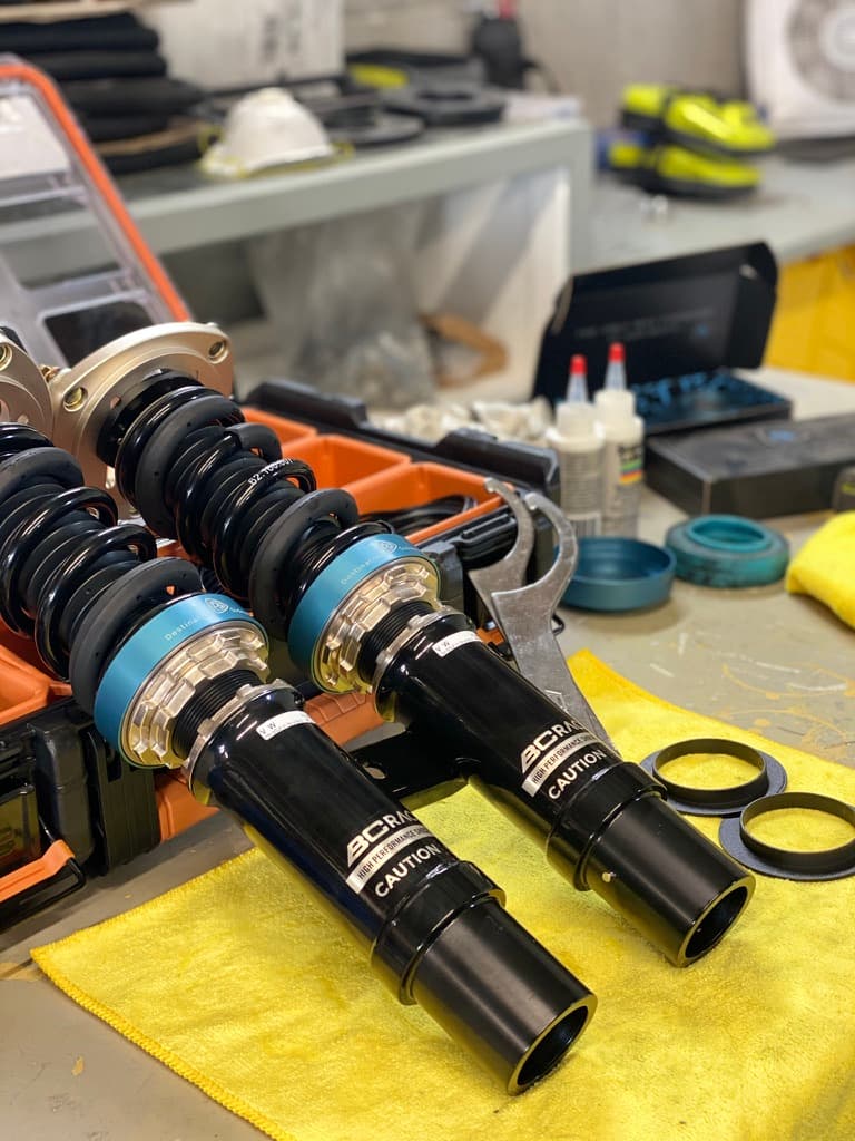 Two BC Racing coilovers on a workbench with blue V1 adapters fitted under the bottom of the springs.