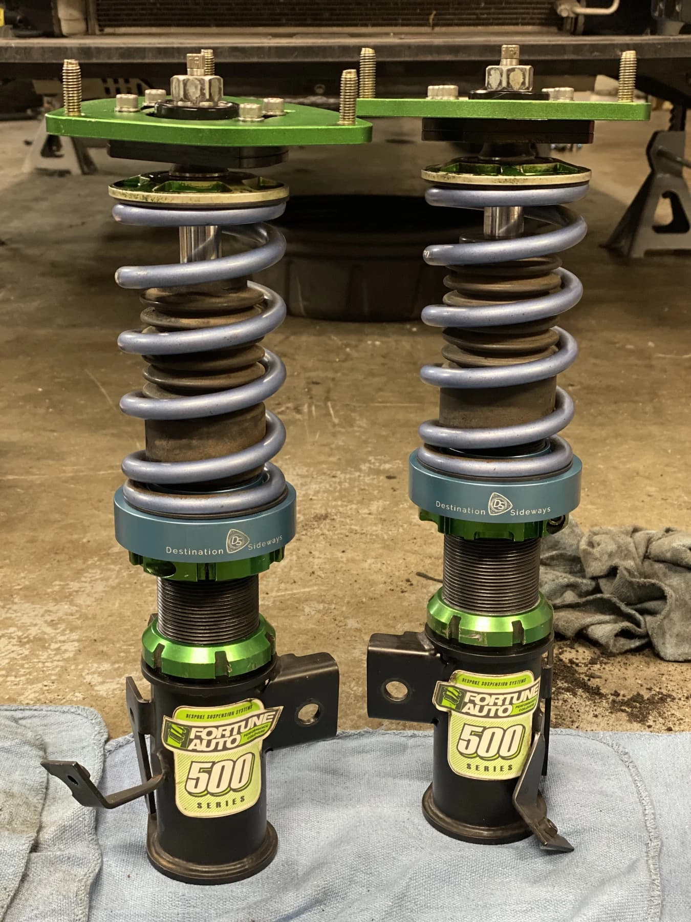 Two coilovers with blue adapters beneath their grey springs on a workbench.